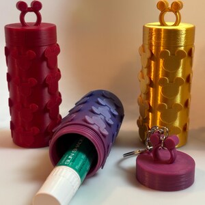 May include: Three cylindrical containers with raised mouse-ear silhouettes on the exterior. One is dark red, one is gold, and one is purple. The purple container is open, revealing a white lip balm tube inside, with its matching purple lid nearby.