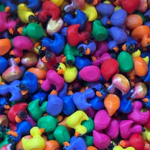 May include: A large collection of small, multi-colored rubber ducks in various shades of yellow, blue, red, green, orange, pink, and purple. Each duck features a distinct beak and eye detail, creating a dense, vibrant pile of toy figures.