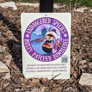 May include: A white sign with a purple circle containing a colorful illustration of a rusty patched bumblebee. The sign reads "Endangered Species * Rusty Patched Bumblebee *" and "This habitat has been planted with native plants specifically for conservation efforts to support the federally endangered rusty patched bumblebee. Design by DreamWeaver Kreati".