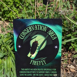 May include: A black and green metal sign with the words "Conservation Area" and "Firefly" in white lettering. The sign features a cartoon image of a firefly with a glowing tail. The text below the image reads "This habitat has been created specifically for conservation efforts to support firefly species. Leave the leaves - dim the lights - watch them glow. Design by Drean Weaver Kreations."