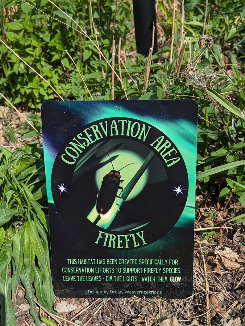 May include: A black and green metal sign with the words "Conservation Area" and "Firefly" in white lettering. The sign features a cartoon image of a firefly with a glowing tail. The text below the image reads "This habitat has been created specifically for conservation efforts to support firefly species. Leave the leaves - dim the lights - watch them glow. Design by Drean Weaver Kreations."
