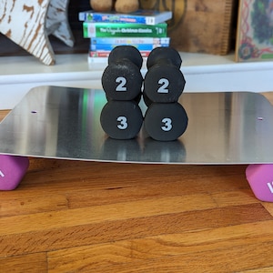 Op de afbeelding: Een set van zes zwarte dumbbells met witte nummers op een zilveren metalen platform. De dumbbells wegen 2, 2, 3, 3, 5 en 5 pond. Het platform staat op een houten vloer.