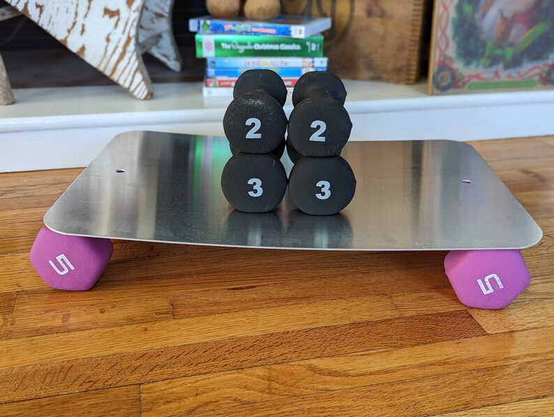 Op de afbeelding: Een set van zes zwarte dumbbells met witte nummers op een zilveren metalen platform. De dumbbells wegen 2, 2, 3, 3, 5 en 5 pond. Het platform staat op een houten vloer.