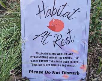 以下が含まれることがあります： 紫とオレンジの水彩画の背景を持つ白い看板。看板には「Habitat At Rest」と書かれており、「At」の文字の上に寝ている漫画のキツネが描かれている。看板には、「この庭では、花粉媒介者と野生動物が冬眠しています。植物は、彼らが冬を乗り切るために必要な避難所を提供しています。邪魔しないでください。」とも書かれている。看板は、茶色の土と緑の草のベッドの上に置かれている。