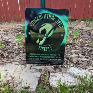 May include: A green and black sign with the words "Conservation Area" and "Firefly" in white. The sign also includes the text "This habitat has been created specifically for conservation efforts to support firefly species. Leave the leaves - dim the lights - watch them glow. Design by Dream Weaver Kreations."