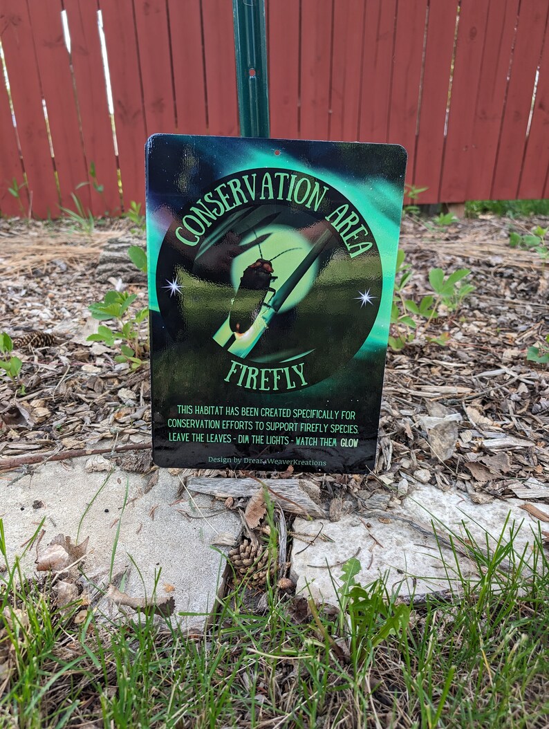 May include: A green and black sign with the words "Conservation Area" and "Firefly" in white. The sign also includes the text "This habitat has been created specifically for conservation efforts to support firefly species. Leave the leaves - dim the lights - watch them glow. Design by Dream Weaver Kreations."