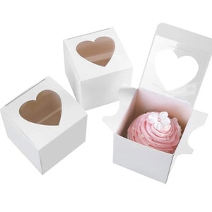 May include: Three white cardboard cupcake boxes with a heart-shaped window. The boxes are designed to hold cupcakes and have a clear window to show off the cupcake inside. One box has a pink cupcake with white frosting and a white flower on top.