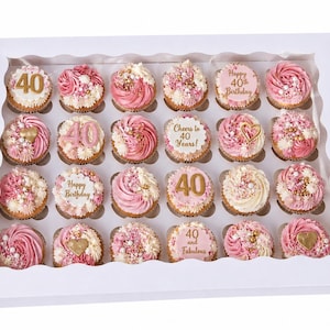 Pode incluir: Uma caixa branca cheia de 24 cupcakes, cada um com cobertura rosa e coberturas decorativas. Alguns cupcakes têm números dourados ou o texto "Happy 40th Birthday" e "Cheers to 40 Years!" Uma sobremesa festiva.