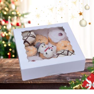 May include: A white cardboard box with a clear window, filled with assorted donuts. The donuts are decorated with white frosting, chocolate, and sprinkles. The box is sitting on a wooden surface.