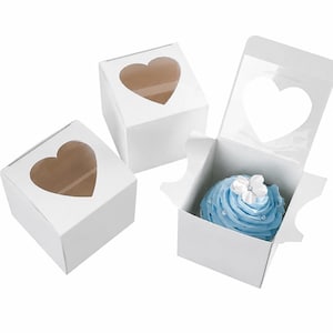 May include: White cupcake boxes with heart-shaped windows. One box is open, revealing a blue frosted cupcake with a white flower and pearl sprinkles. Two other boxes are closed.
