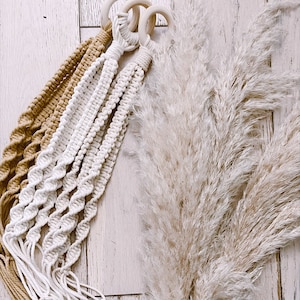 May include: Two macrame plant hangers with wooden rings, one in beige and one in white, hanging against a white wood background with a dried pampas grass bouquet.