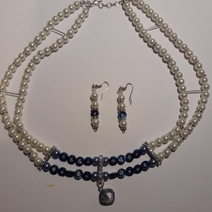 May include: A two-strand necklace and matching earrings set. The necklace features white and navy blue beads, with a silver pendant and accents. The earrings have white and navy blue beads. The jewelry set is on a white background.