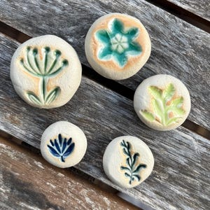 Puede incluir: Cinco botones de cerámica circulares con un acabado mate en color blanco roto, cada uno con un diseño botánico en relieve único. Los patrones grabados están resaltados con esmaltes en tonos turquesa, verde claro, azul oscuro y verde salvia.