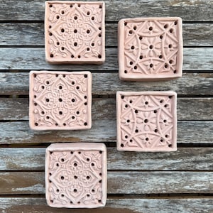 Può includere: Cinque piastrelle quadrate in terracotta rosa chiaro con intricati motivi in rilievo e piccoli fori circolari. Ogni piastrella presenta un design decorativo simmetrico con linee curve e motivi di ispirazione floreale.