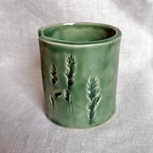 Könnte beinhalten: Eine handgefertigte, zylindrische Keramiktasse in einem gedämpften Grünton. Die Tasse hat ein geprägtes Design aus hohem Gras oder Weizenähren. Das Design ist mit dunkelgrünen Linien detailliert. Die Tasse hat eine glatte, glänzende Oberfläche.