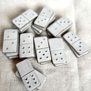 Puede incluir: Un conjunto de dominós grises claros con puntos blancos, dispuestos sobre una tela de tono neutro. Los dominós están apilados y dispersos, mostrando su forma rectangular y los patrones de puntos.