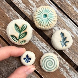 Könnte beinhalten: Eine Sammlung von fünf kleinen, runden Keramiksteinen mit verschiedenen geprägten Designs. Die Designs umfassen ein Blatt, eine Spirale, eine Fleur-de-Lis und andere botanische Muster, alle in Grün- und Blautönen. Die Steine sind cremefarben und haben einen Durchmesser von etwa 2,5 cm.