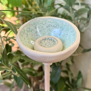 Könnte beinhalten: Ein Keramik-Gartenstecker mit einer flachen, schalenförmigen Oberseite, die außen matt cremeweiß und innen glänzend mintgrün gesprenkelt ist. In der Mitte der Schale befindet sich ein kleines, rundes, strukturiertes Blumenmedaillon auf einem Stab.