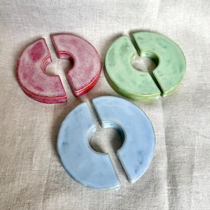 Könnte beinhalten: Drei Weinglas-Anhänger aus Keramik in Rot-, Grün- und Blautönen. Jeder Anhänger ist ein geteilter Kreis mit einem zentralen Loch, der zum Anklemmen an den Stiel eines Weinglases bestimmt ist. Die Anhänger sind auf einer neutralen Leinenoberfläche angeordnet.