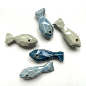 Op de afbeelding: Vijf keramische visornamenten in tinten blauw en grijs. De vissen zijn geometrisch van vorm en hebben een gat aan de zijkant.