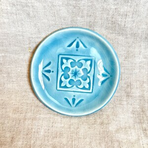 Può includere: Un piccolo piatto rotondo in ceramica azzurra con un motivo floreale bianco decorativo al centro. Il piatto ha una glassa craquelé e misura circa 10 cm di diametro. Il design include frecce stilizzate e motivi floreali.