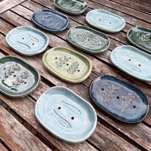 Può includere: Una collezione di portasapone in ceramica in varie tonalità di blu, verde e verde acqua. Ogni piatto ovale presenta disegni botanici o di pesci in rilievo e fori di drenaggio. I piatti sono disposti su una superficie di legno.