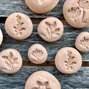 Op de afbeelding: Een verzameling van tien ronde, lichtbruine kleischijven, elk met een uniek in reliëf gemaakt botanisch ontwerp. De texturen omvatten diverse bloemmotieven, bladertakjes en ingewikkelde plantmotieven die in het oppervlak van de gladde, mat afgewerkte kleistukken zijn gedrukt.