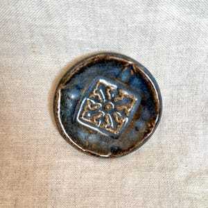 Pode incluir: Uma base de cerâmica artesanal com um esmalte azul escuro e um desenho quadrado em relevo no centro. A base tem uma forma rústica e orgânica e uma borda marrom. O desenho no centro é um padrão floral estilizado.