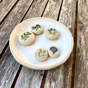 Könnte beinhalten: Eine runde, weiße Keramikschale mit fünf kleinen, kreisförmigen, cremefarbenen Keramikplättchen. Jedes Plättchen zeigt ein einzigartiges, vertieftes Pflanzen- oder Blumenmotiv, das in Blau-, Grün- und Orangetönen glasiert ist und eine Textur aufweist.