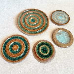 Può includere: Un set di cinque sottobicchieri in ceramica con disegni a cerchi concentrici. I sottobicchieri presentano una combinazione di colori marrone, verde acqua e azzurro. Di varie dimensioni, hanno una finitura lucida, adatti per l'arredamento.
