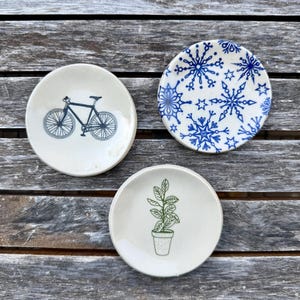 Può includere: Tre piccoli piatti rotondi in ceramica color bianco sporco. Uno presenta un'illustrazione di una bicicletta blu scuro, un altro un motivo a fiocchi di neve blu e il terzo un disegno di una pianta in vaso verde. Finitura smaltata e liscia.