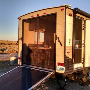 May include: A tan and brown camper trailer with a screen door open, revealing a table and chairs inside. The trailer has a ramp and a step, and the text "GREY WOLF" is visible on the side.