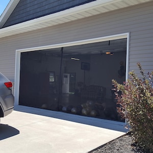 Op de afbeelding: Een grijs huis met een zwarte intrekbare screendeur die over een garagedeur is geïnstalleerd. De screendeur staat open en de garage is zichtbaar door het scherm. Er zijn verschillende ronde objecten in de garage, mogelijk ballen of trainingsapparatuur.