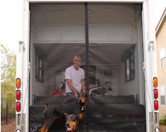 May include: Two black Doberman Pinschers stand in front of a white camper van with the rear doors open. The van has a black mesh screen door fitted. The dogs are looking at the camera.