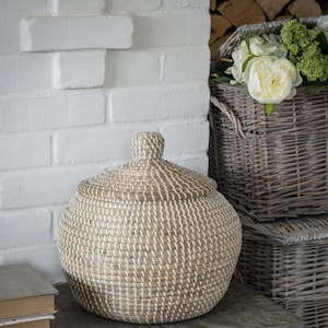 May include: A round, lidded basket with a woven design in natural and white tones. The basket has a knob on top of the lid. A stack of books and a white brick wall are in the background. Other baskets are also visible.