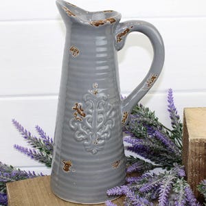 May include: A grey ceramic jug with a handle and an embossed decorative design. The jug has a distressed finish with brown accents. The jug is on a wooden surface with purple lavender sprigs.