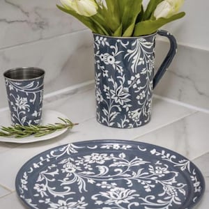 May include: A dark blue metal pitcher, a matching tumbler, and a round tray, all featuring a white floral pattern. The pitcher is filled with white artificial tulips. The set displays a consistent botanical design across each individual piece.