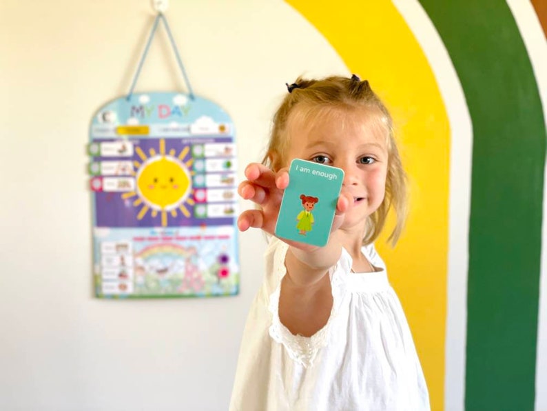 May include: A young person holds up a blue affirmation card with the text "I am enough" and a cartoon illustration of a girl. The card is in front of a colorful rainbow wall.