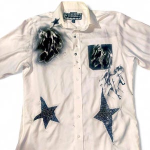 May include: A white button-down shirt featuring black airbrushed graphics of winged figures, a horse, and stars. The shirt includes a chest pocket, a collar, and a label at the neck that reads "stellamakes". Two large star-shaped fabric patches are stitched onto the lower front panels.
