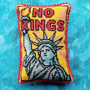 Puede incluir: Un pequeño cojín rectangular con un diseño de punto de cruz que presenta la Estatua de la Libertad y las palabras "NO KINGS" en letras rojas. El cojín tiene un fondo amarillo, un borde rojo con pompones y está sobre un fondo azul.