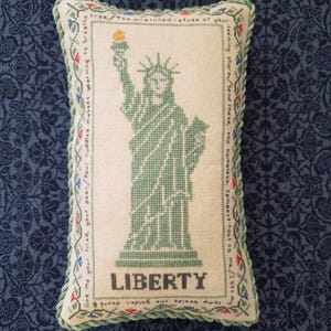 May include: A rectangular decorative pillow featuring a cross-stitch design of the Statue of Liberty in green. The word "LIBERTY" is stitched below the statue. The pillow has a green and blue rope trim and floral border with text.