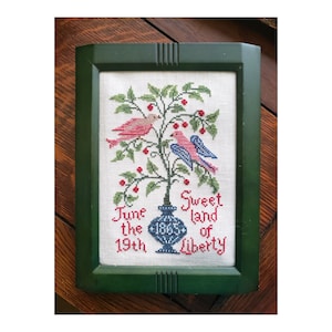 May include: A framed cross-stitch embroidery depicting a vase with a flowering plant and three birds. The vase is blue and white, the plant has green leaves and red berries, and the birds are pink and blue. The text reads "June the 19th 1865 Sweet land of Liberty".