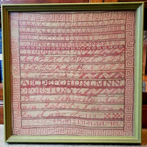 May include: A framed sampler with a green frame and a cream-colored background. The sampler features a red and green alphabet, a red and green number line, and the text "Par la vertu de l'ame" and "Em. Ed. Jan. 1842".