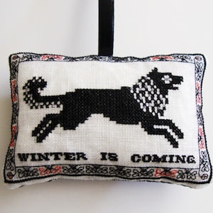 May include: A white pillow with a black embroidered direwolf and the text "Winter is Coming". The pillow has a black and red border.