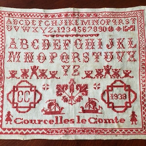 May include: A white linen sampler with red embroidery. The sampler includes the alphabet, numbers, and a variety of designs, including a ship, a flower, and a monogram. The sampler is dated 1938 and signed "Courcelles le Comte".