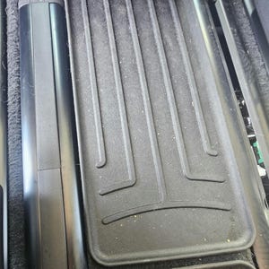 May include: A black automotive floor mat featuring a textured surface with raised vertical channels and a curved horizontal border, positioned alongside a smooth black plastic trim piece within a vehicle interior floor space.