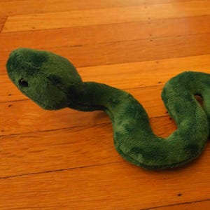 May include: A plush toy shaped like a snake, covered in soft, dark green fabric. The snake has a rounded head with a visible black eye and a long, winding body that curves across the frame, featuring a consistent fuzzy texture throughout.