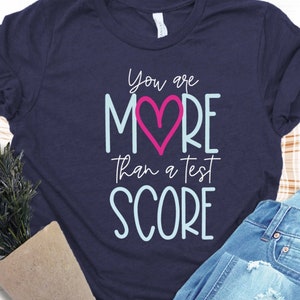 May include: Navy blue t-shirt with the message "You are MORE than a test SCORE" in white, light blue, and pink. The word "MORE" features a pink heart. The shirt is paired with blue jeans.