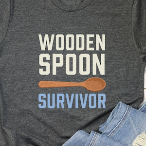 May include: A dark gray t-shirt with the text "WOODEN SPOON SURVIVOR" in white and blue lettering. A brown wooden spoon is pictured above the text.