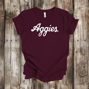 May include: A maroon t-shirt with the word "Aggies" in white script across the front. The shirt is made of a soft material and has rolled-up sleeves. The background is a wooden surface.